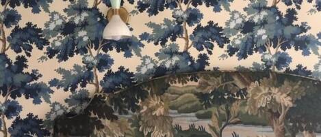 A room with a patterned wallpaper featuring a forest theme, a decorative vase on a table, and a landscape painting on the wall.