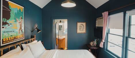 A bedroom with a large bed, a wooden ceiling, a wall-mounted lamp, and a poster of a fly fishing scene.