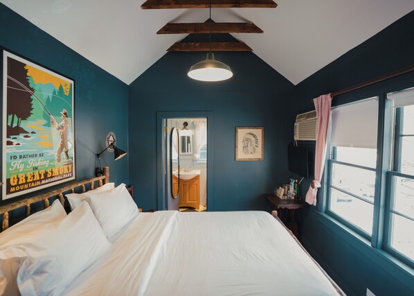 A bedroom with a large bed, a wooden ceiling, a wall-mounted lamp, and a poster of a fly fishing scene.