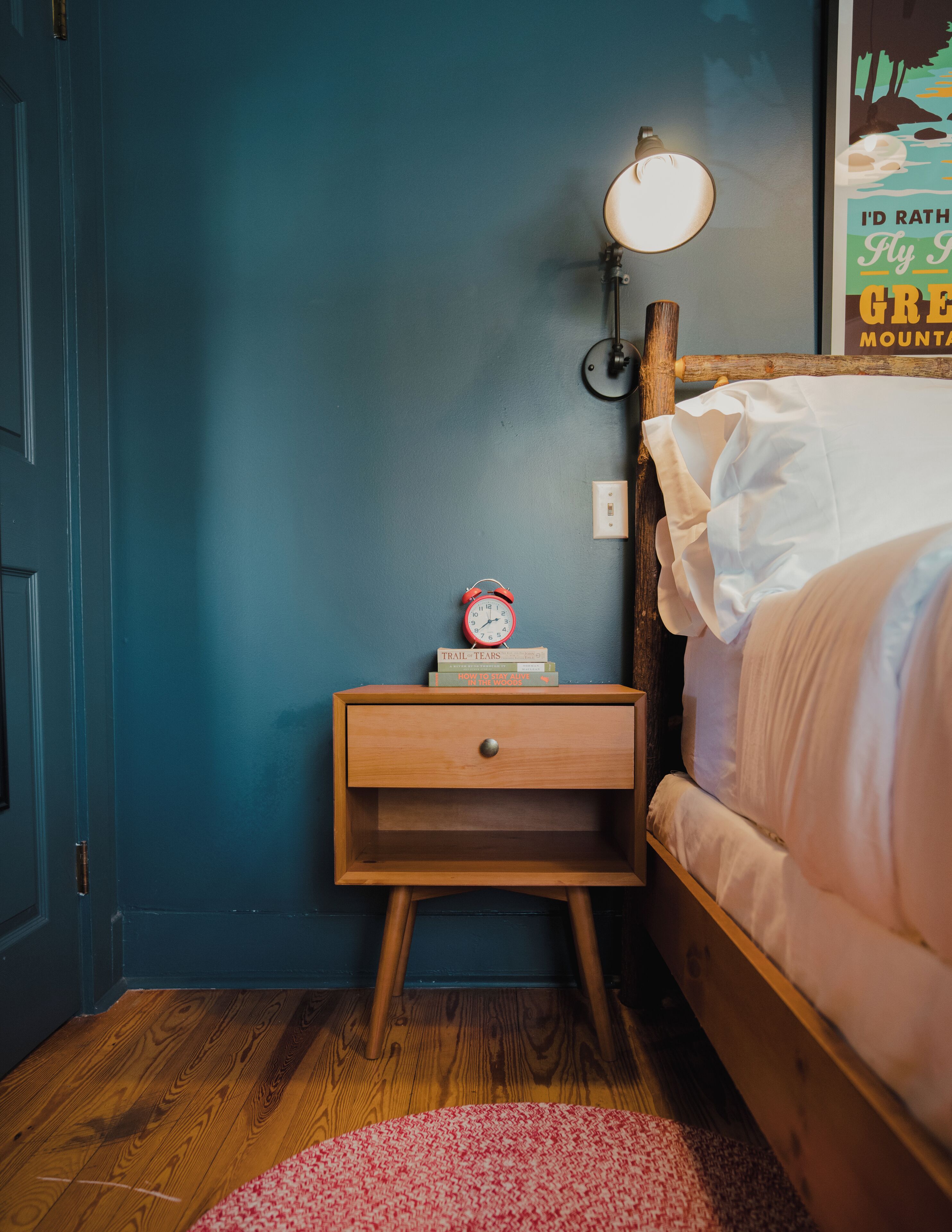 Une chambre avec une table de chevet en bois, une lampe de chevet, un lit avec une literie blanche et un petit tapis.