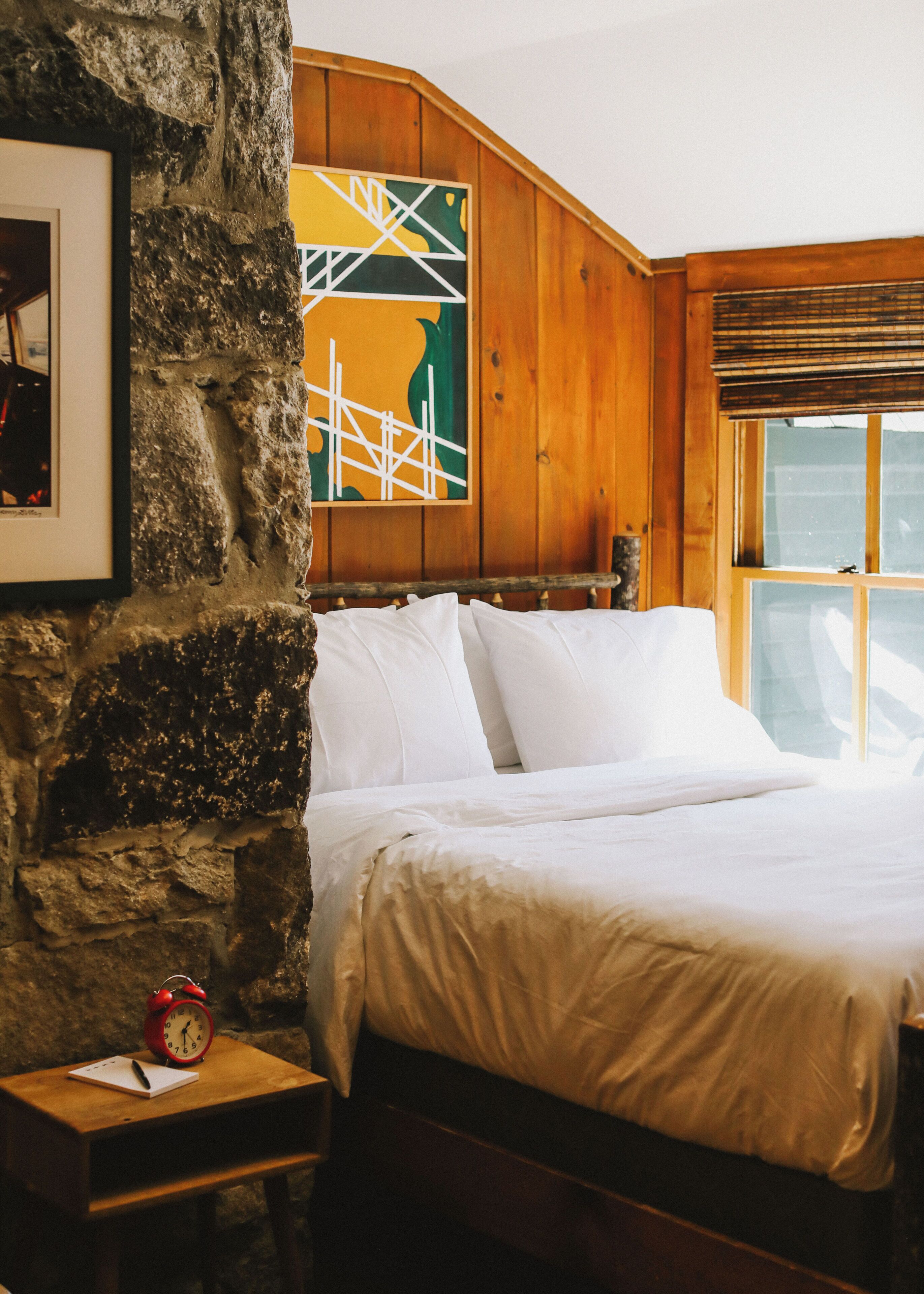 A bedroom with a stone wall, wooden paneling, a bed with white bedding, a nightstand with a red alarm clock, and a framed picture.