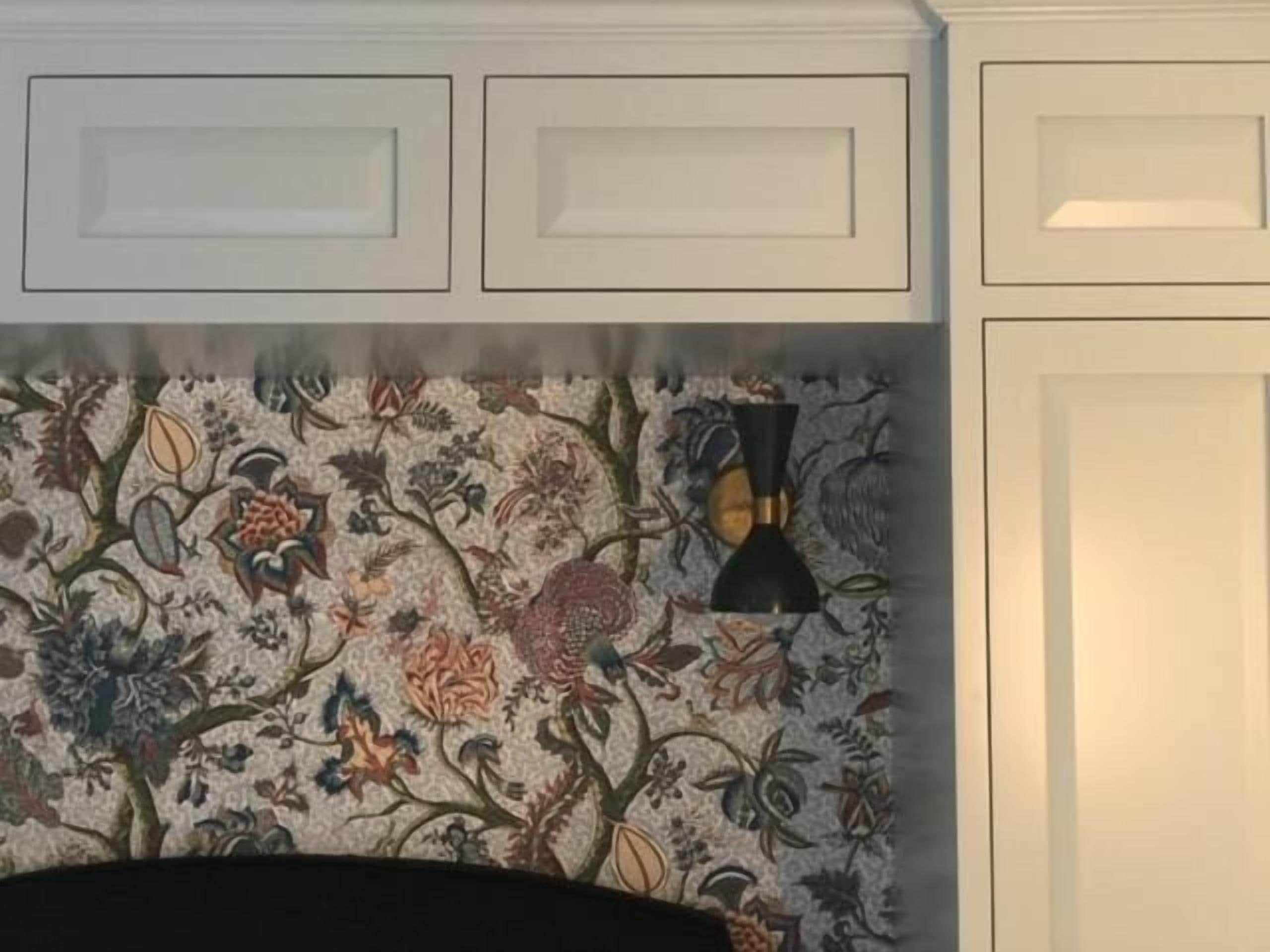 Una habitación con papel tapiz estampado con motivos florales, una lámpara negra y muebles de color blanco.