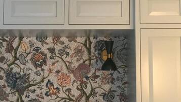 Una habitación con papel tapiz estampado con motivos florales, una lámpara negra y muebles de color blanco.