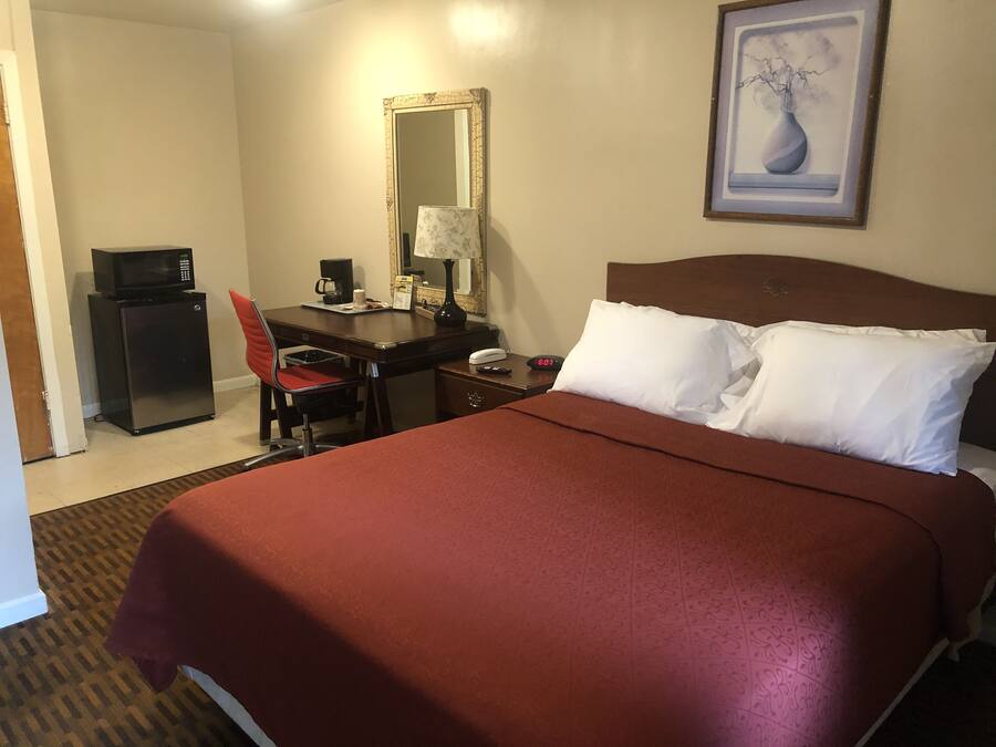 A hotel room with a bed, a desk with a chair, a microwave, a coffee maker, a lamp, a clock, and a framed picture on the wall.