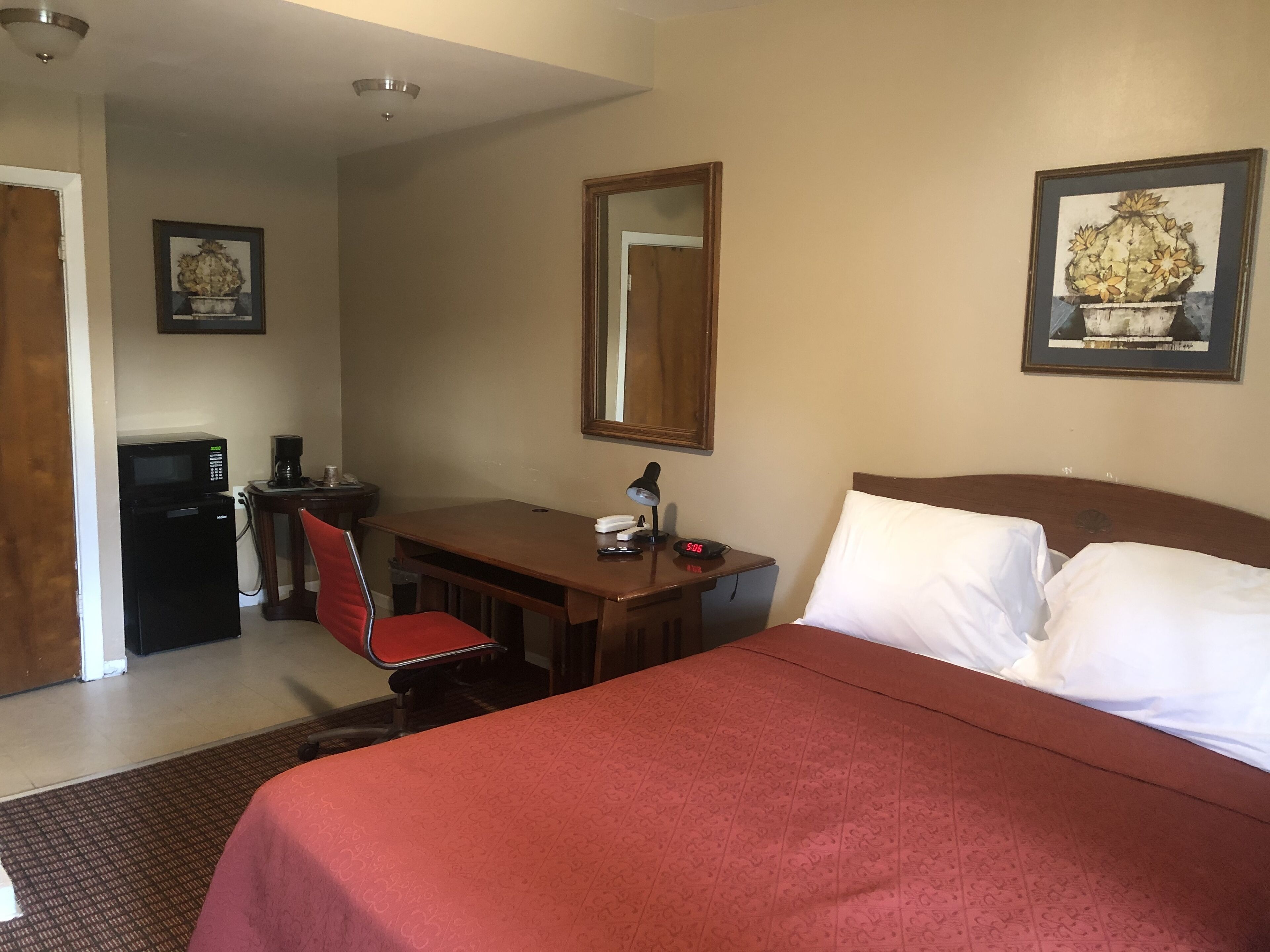 Habitación de hotel con una cama, un escritorio con silla, microondas, cafetera, reloj y un cuadro en la pared.