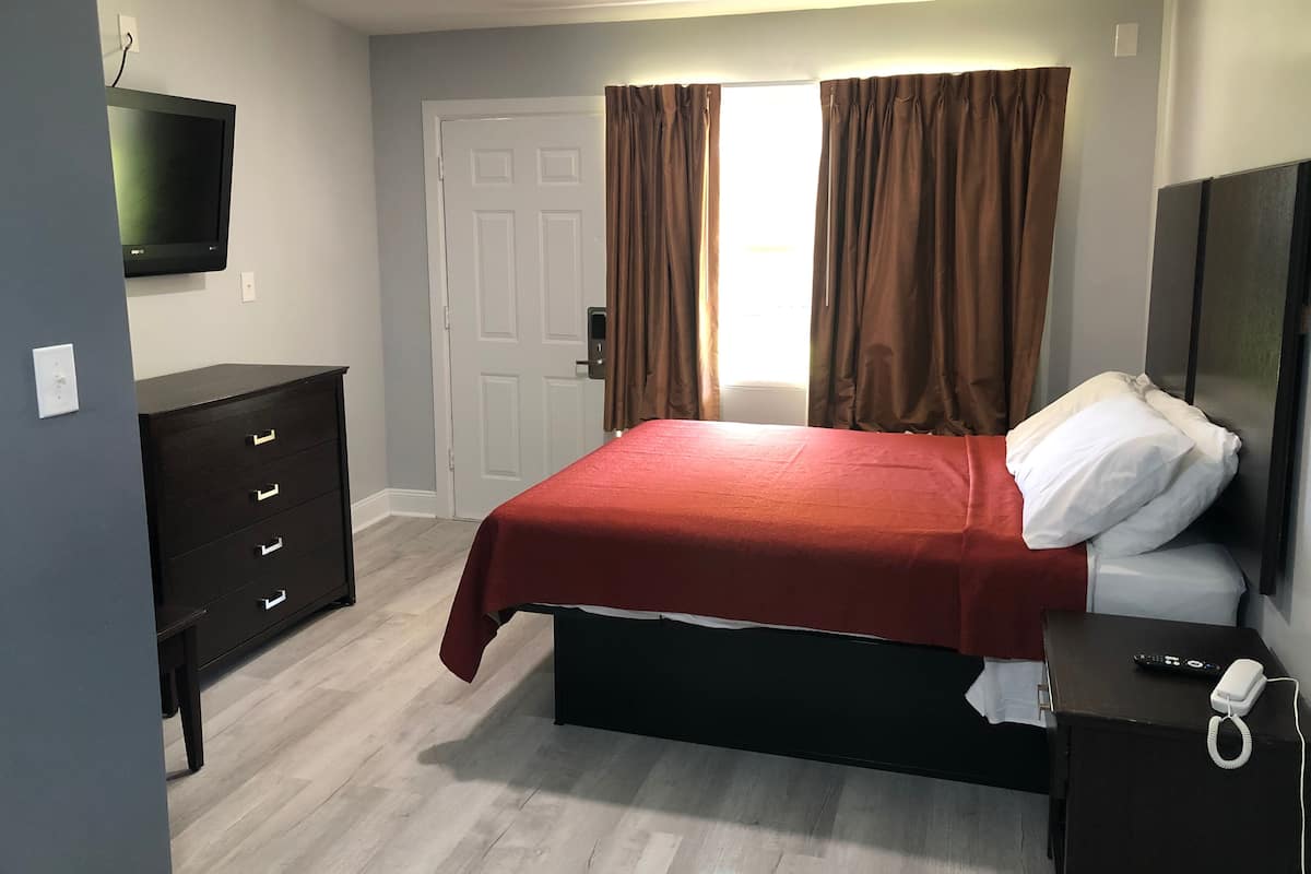 Quarto com cama, cômoda, televisão, porta e janela com cortinas.