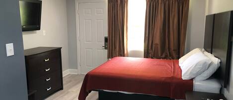 Un dormitorio con cama, cómoda, televisor, puerta y ventana con cortinas.