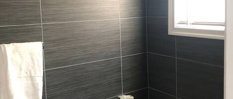 Ein Badezimmer mit dunkel gefliesten Wänden, einer weißen Toilette, einem weißen Handtuch an der Wand und einem Fenster, das für natürliches Licht sorgt.