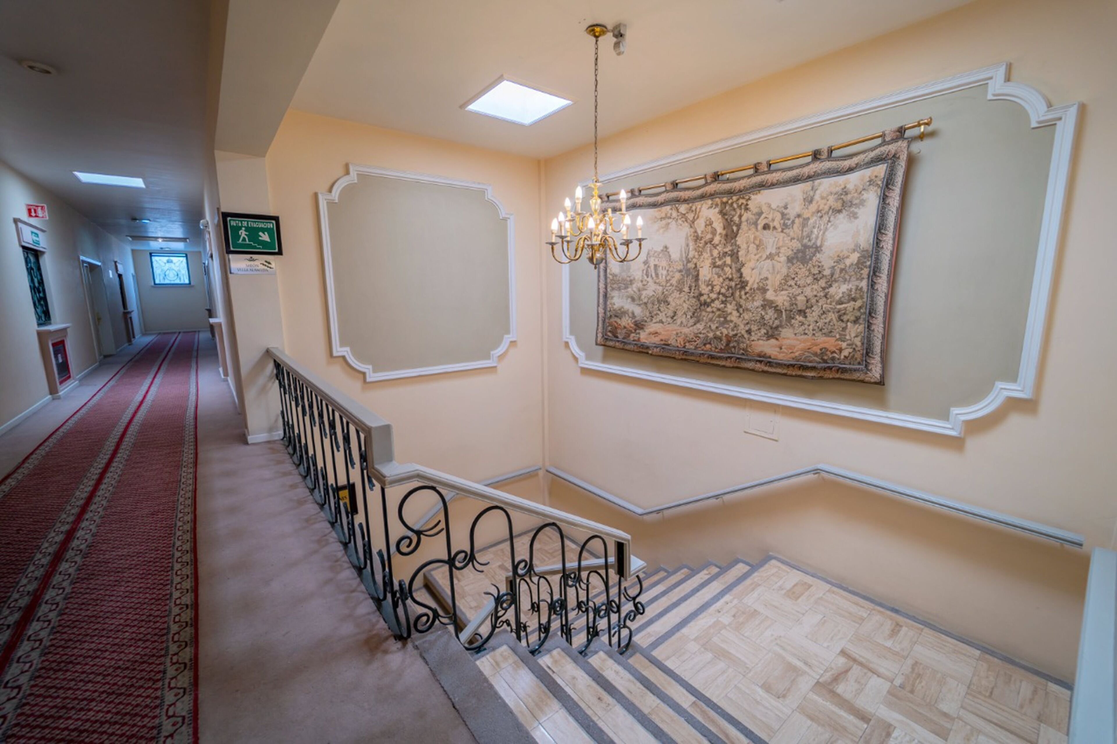 Un couloir avec une moquette rouge, un escalier avec des rampes noires et une grande tapisserie au mur.