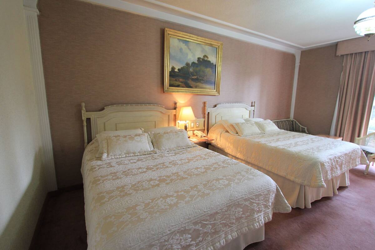 A hotel room with two beds, a painting on the wall, and a lamp on the nightstand.