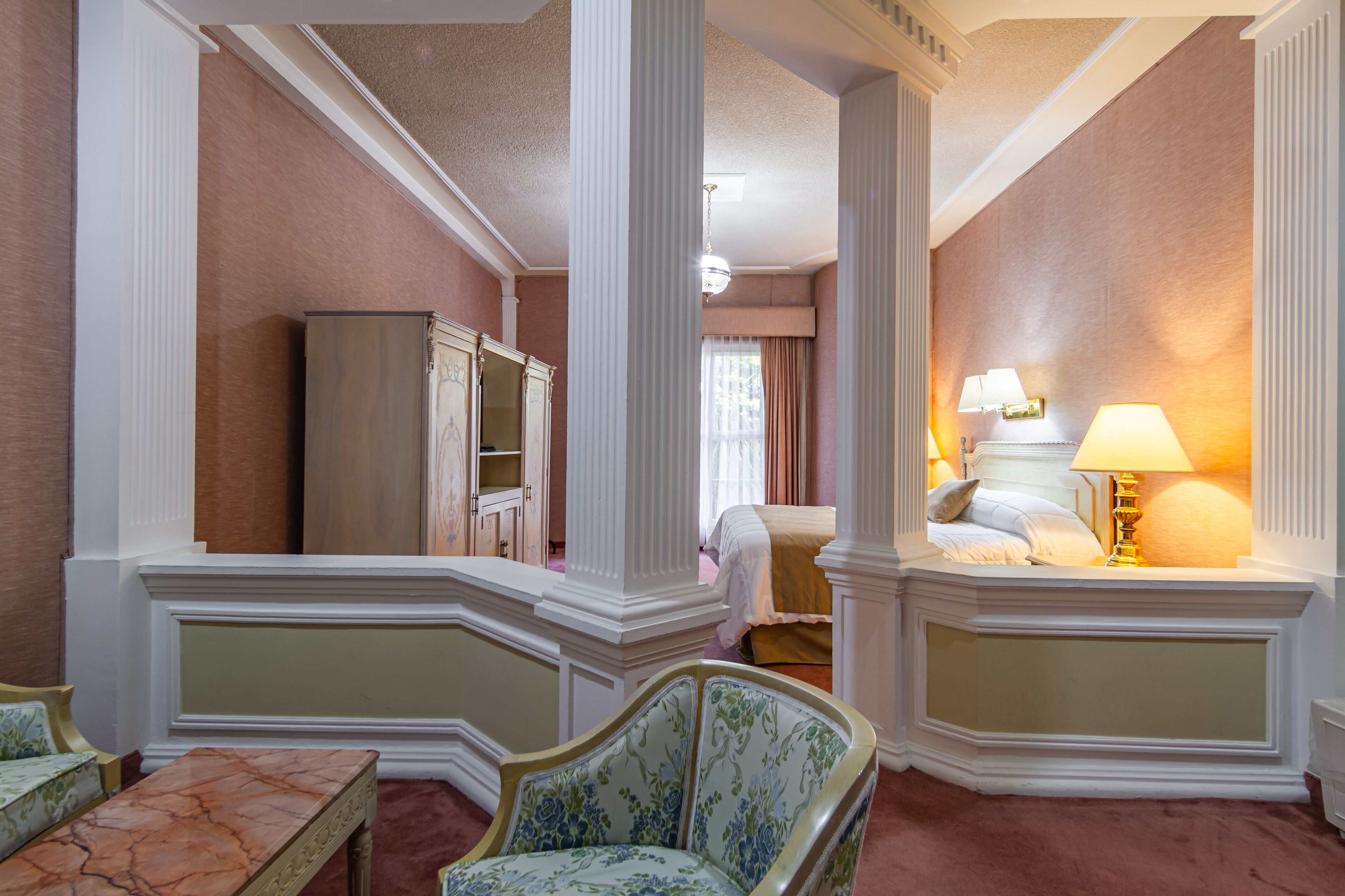 Una habitación de hotel con una mesa de mármol, un sillón con estampado floral, una cama con cabecera blanca y una mesita de noche con lámpara.