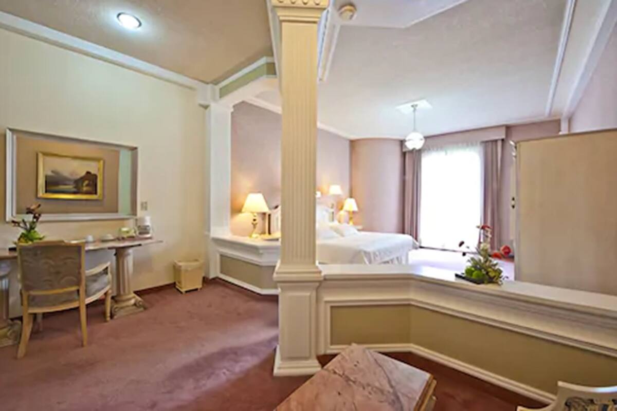 A hotel room with a bed, bedside table, lamp, chair, desk, and a painting on the wall.