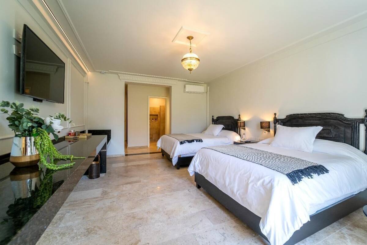 A hotel room with a large bed, a smaller bed, a desk, a TV, and a chandelier.