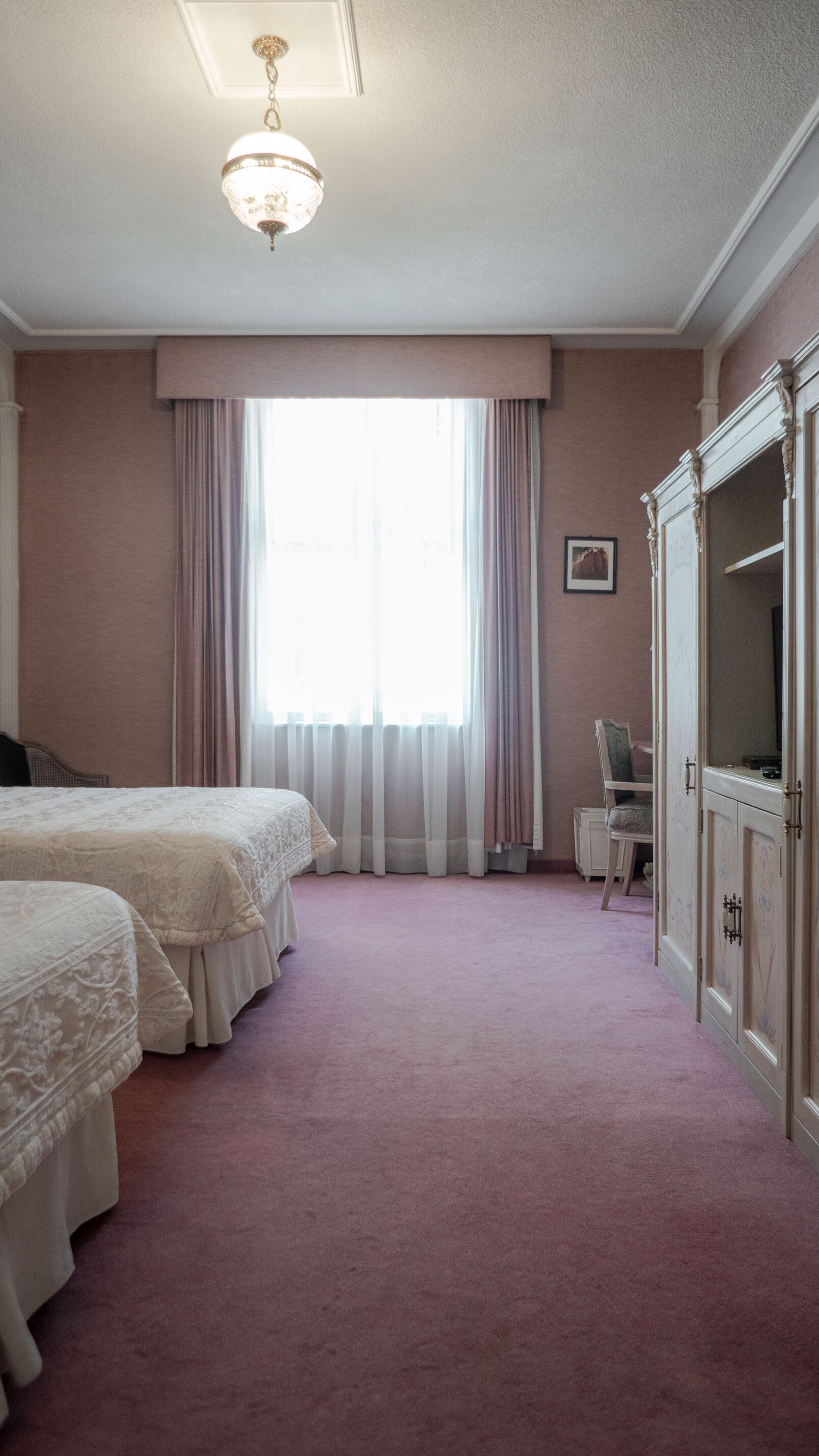 Ett hotellrum med två sängar, ett stort fönster med gardiner, ett skrivbord med en stol och en garderob.