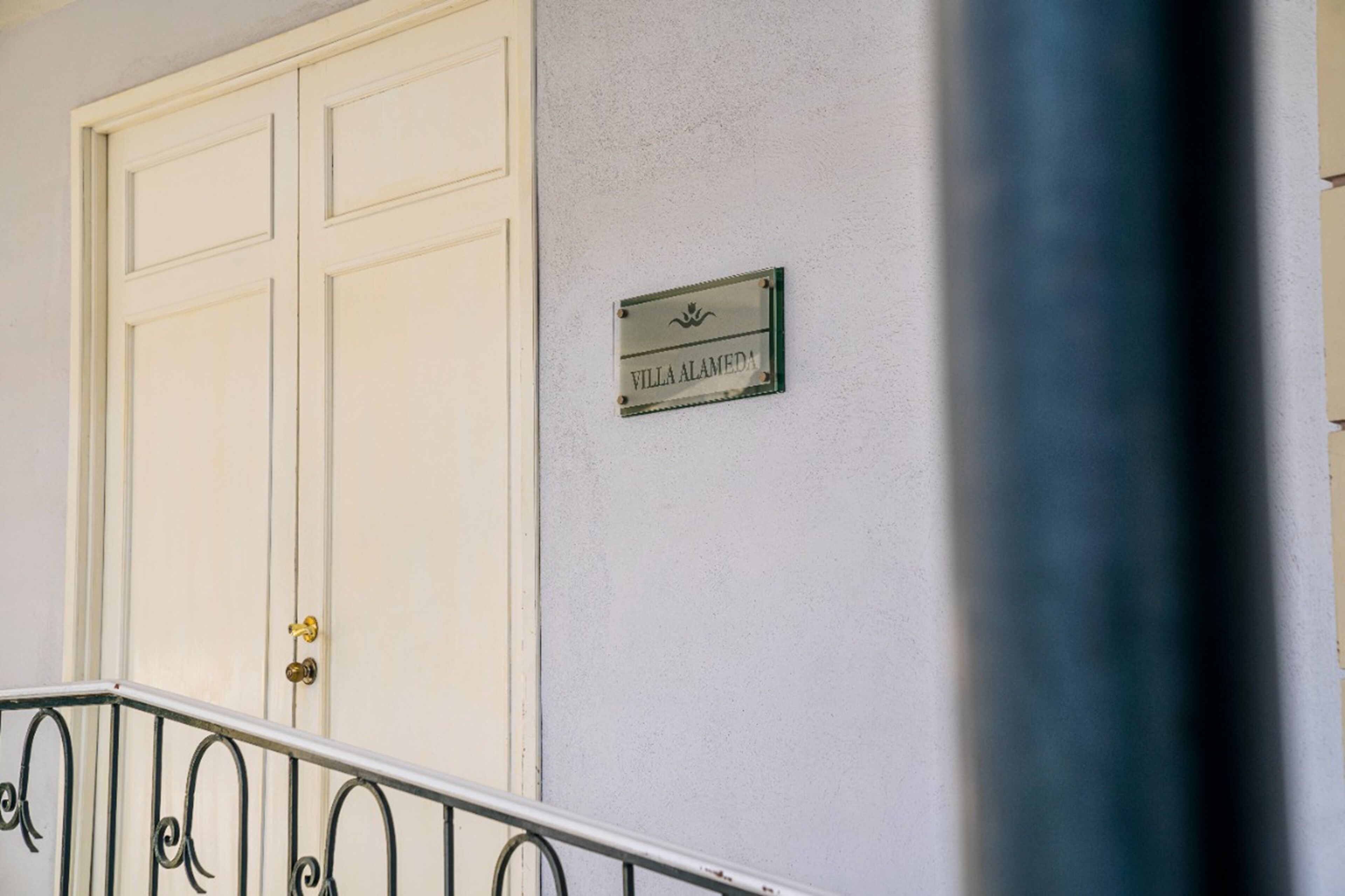 Una puerta blanca con un pomo de latón, una barandilla metálica y un letrero en la pared que dice "Villa Alameda".