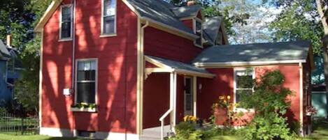 A two-story red house with a front porch, a chimney, and a small garden with flowers.