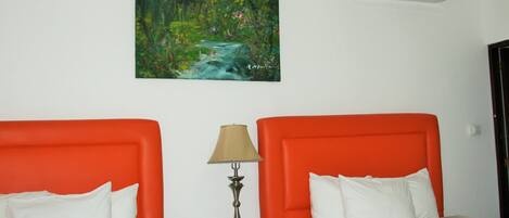 A hotel room with two beds, each with an orange headboard, white bedding, and a painting of a forest stream above the beds.
