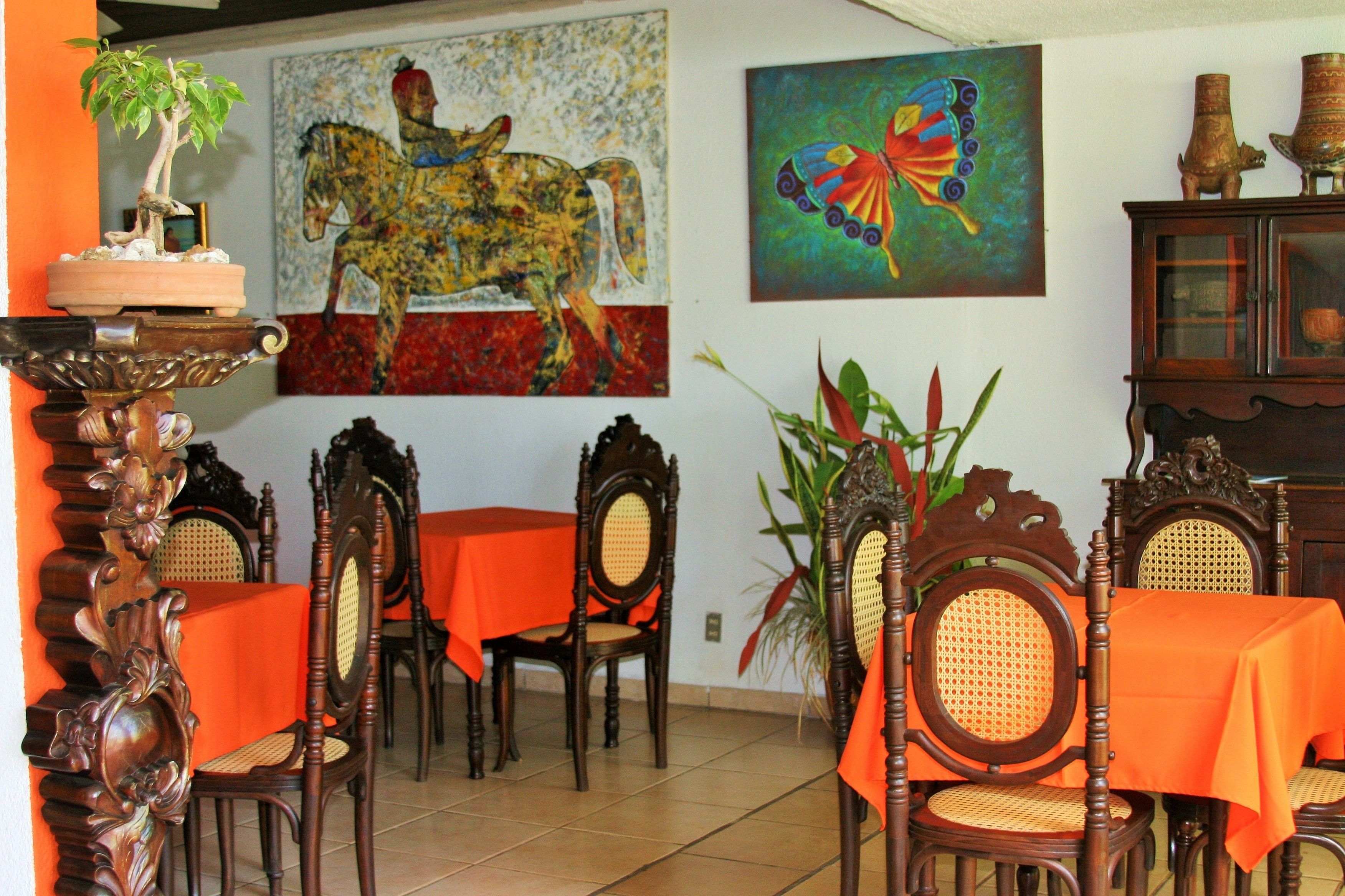 Zona pranzo con sedie e tavoli in legno, una pianta in vaso e due quadri alle pareti.