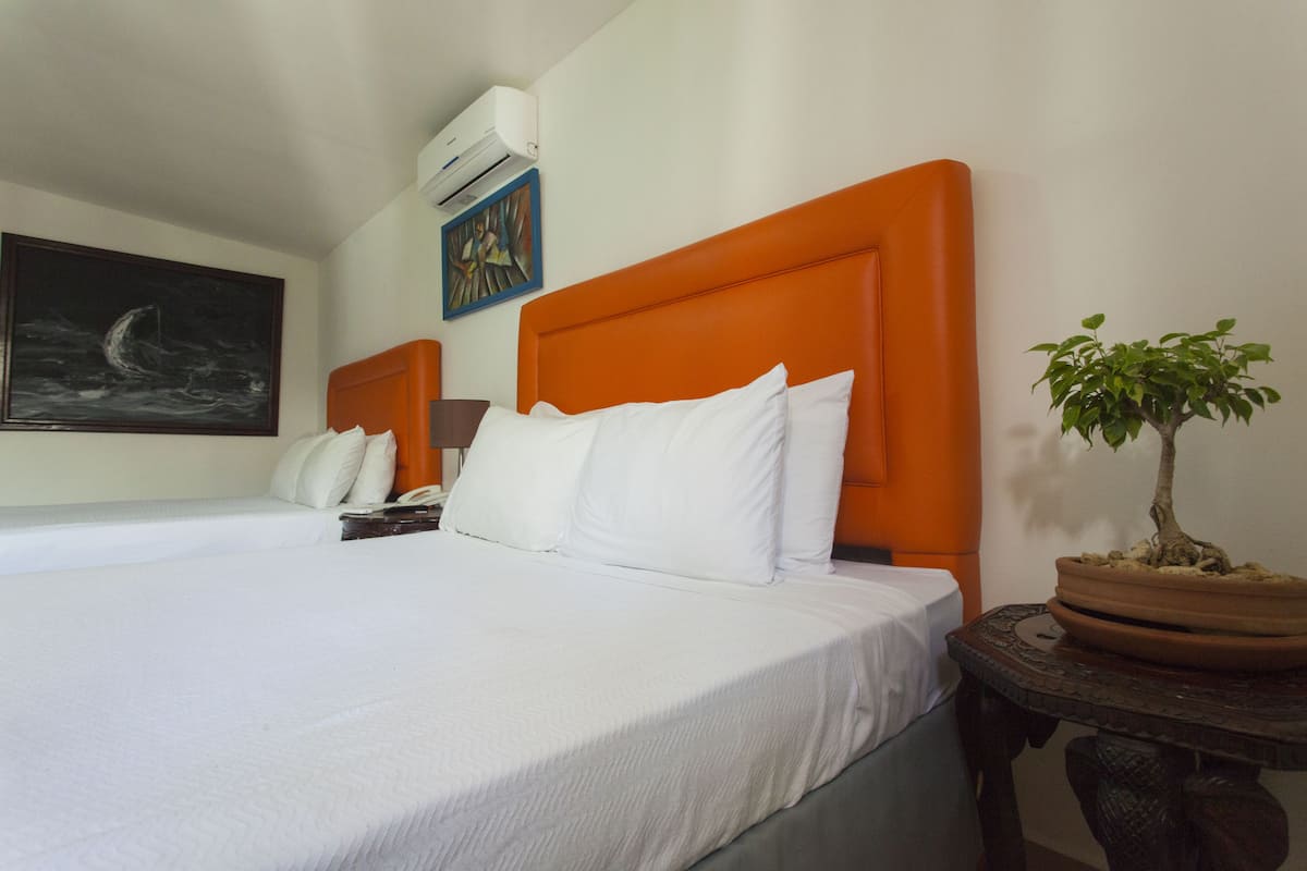 A hotel room with two beds, each with white bedding and orange headboards. There is a potted plant on a small table beside one bed.