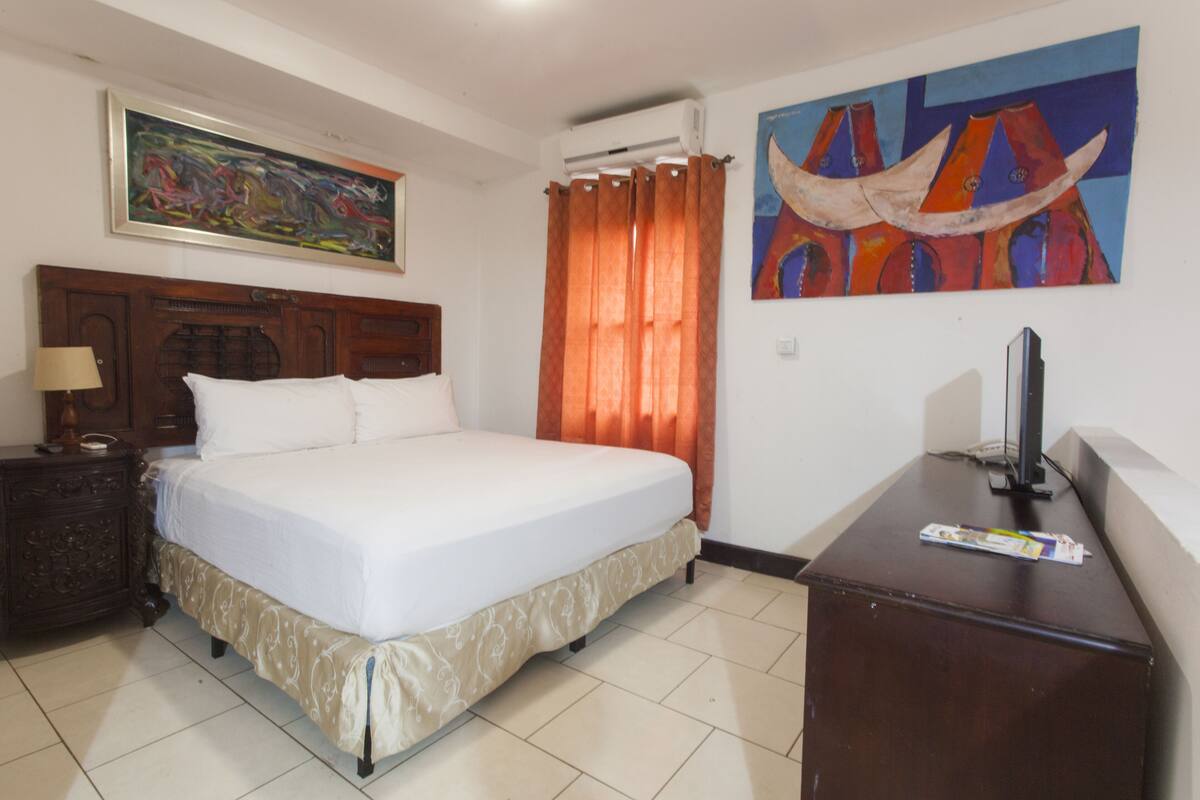 A hotel room with a large bed, a wooden headboard, a nightstand, a desk, a television, and two paintings on the walls.