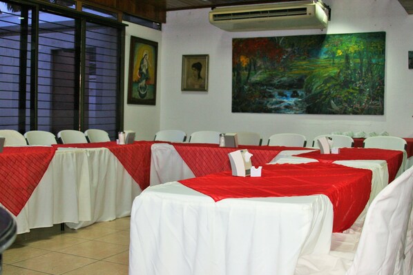 Un salón de conferencias con mesas cubiertas con manteles blancos y rojos, sillas blancas y un cuadro grande en la pared.