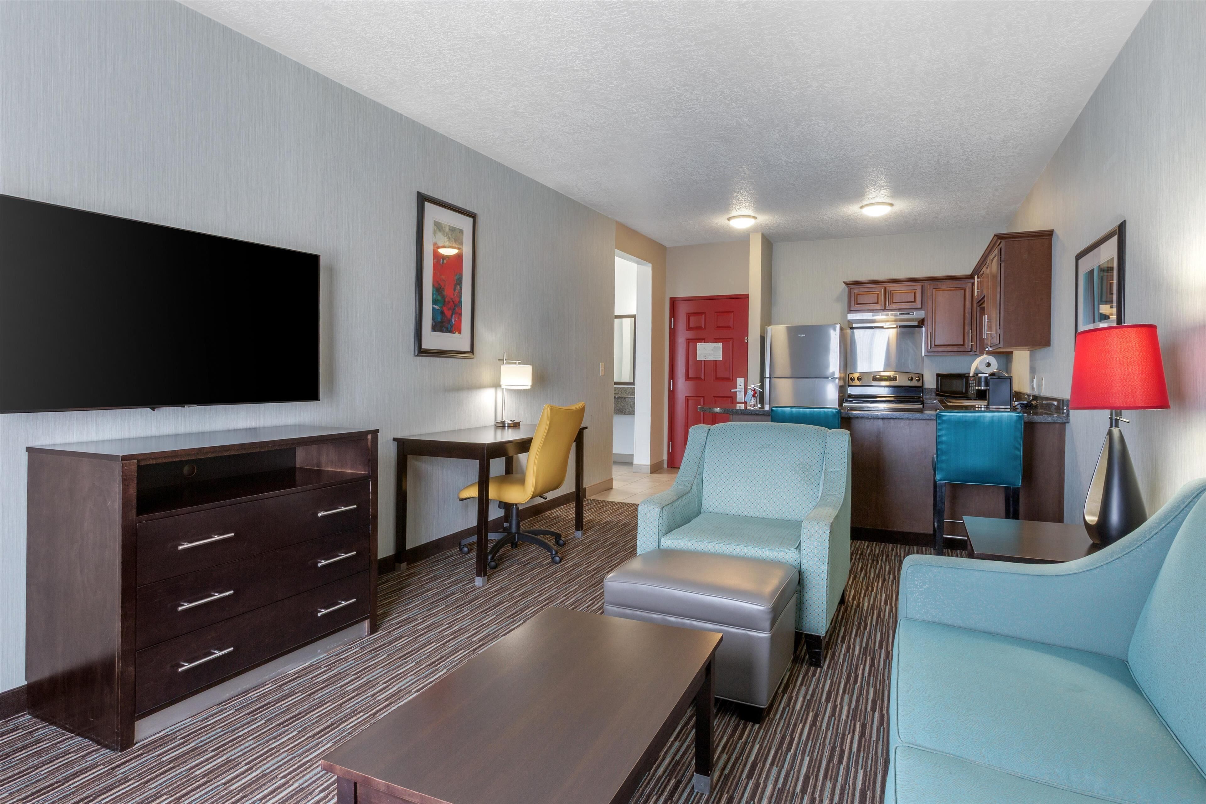 A hotel room with a flat-screen TV, a desk, a chair, a sofa, a kitchen area with a refrigerator, microwave, and stove, and a red door.