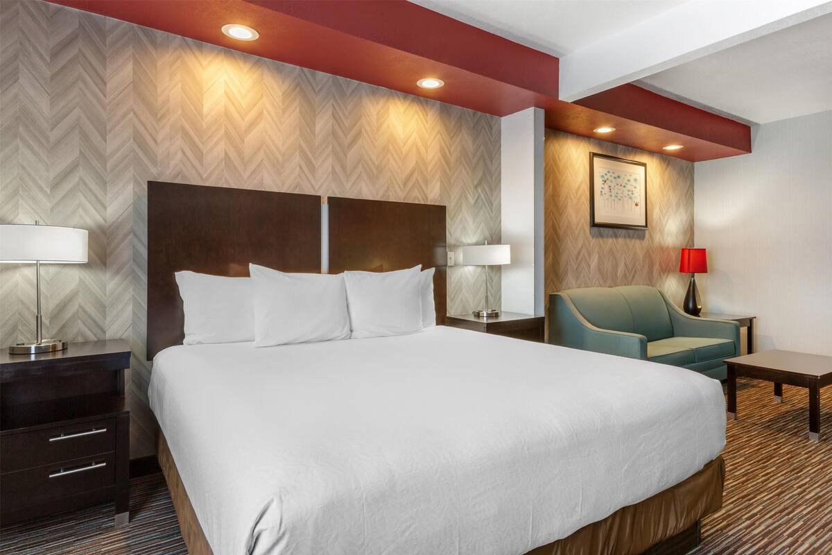 A hotel room with a large bed, a headboard, two bedside tables with lamps, a sofa, and a red lamp on the wall.