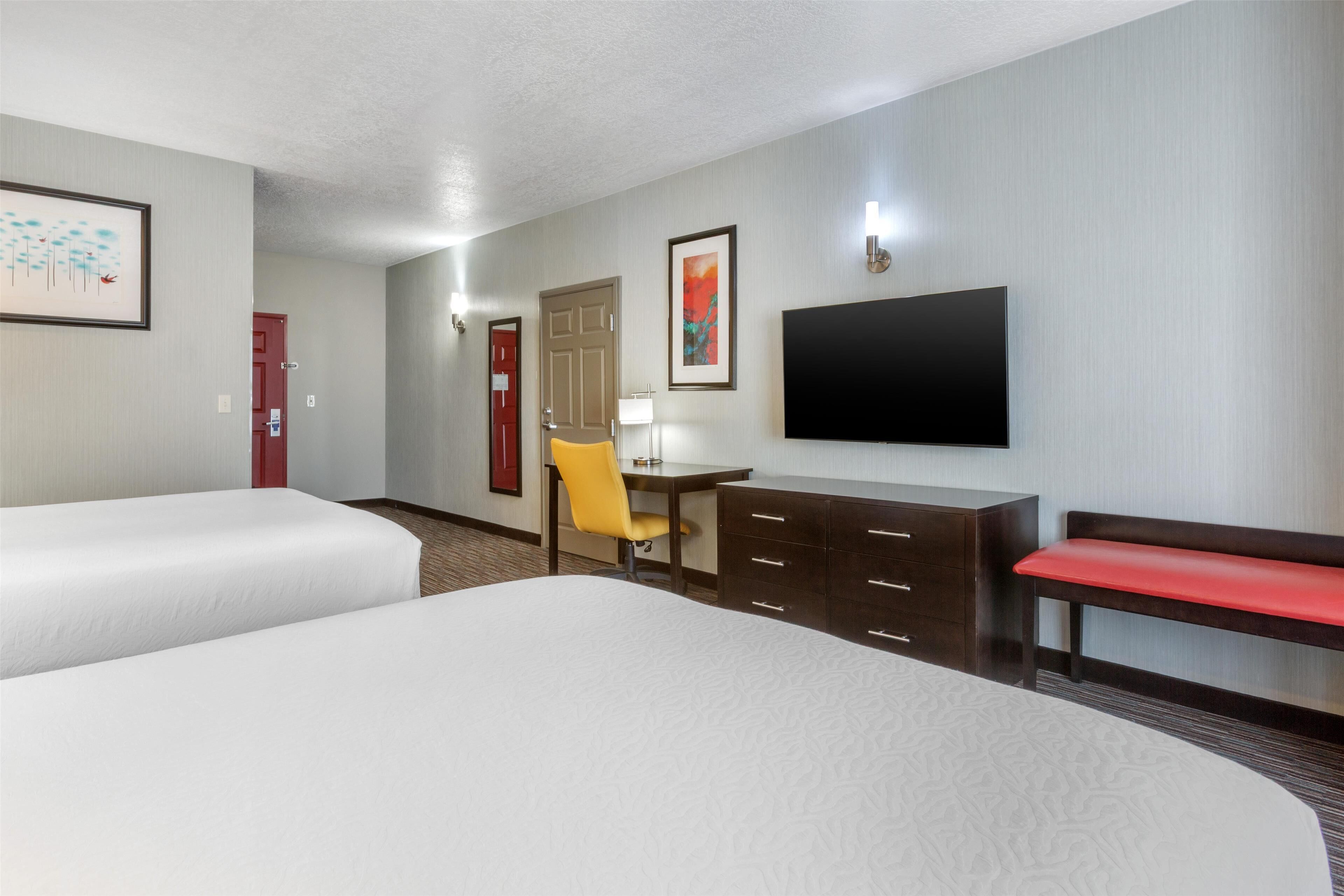 A hotel room with two beds, a desk with a chair, a TV, and a bench.