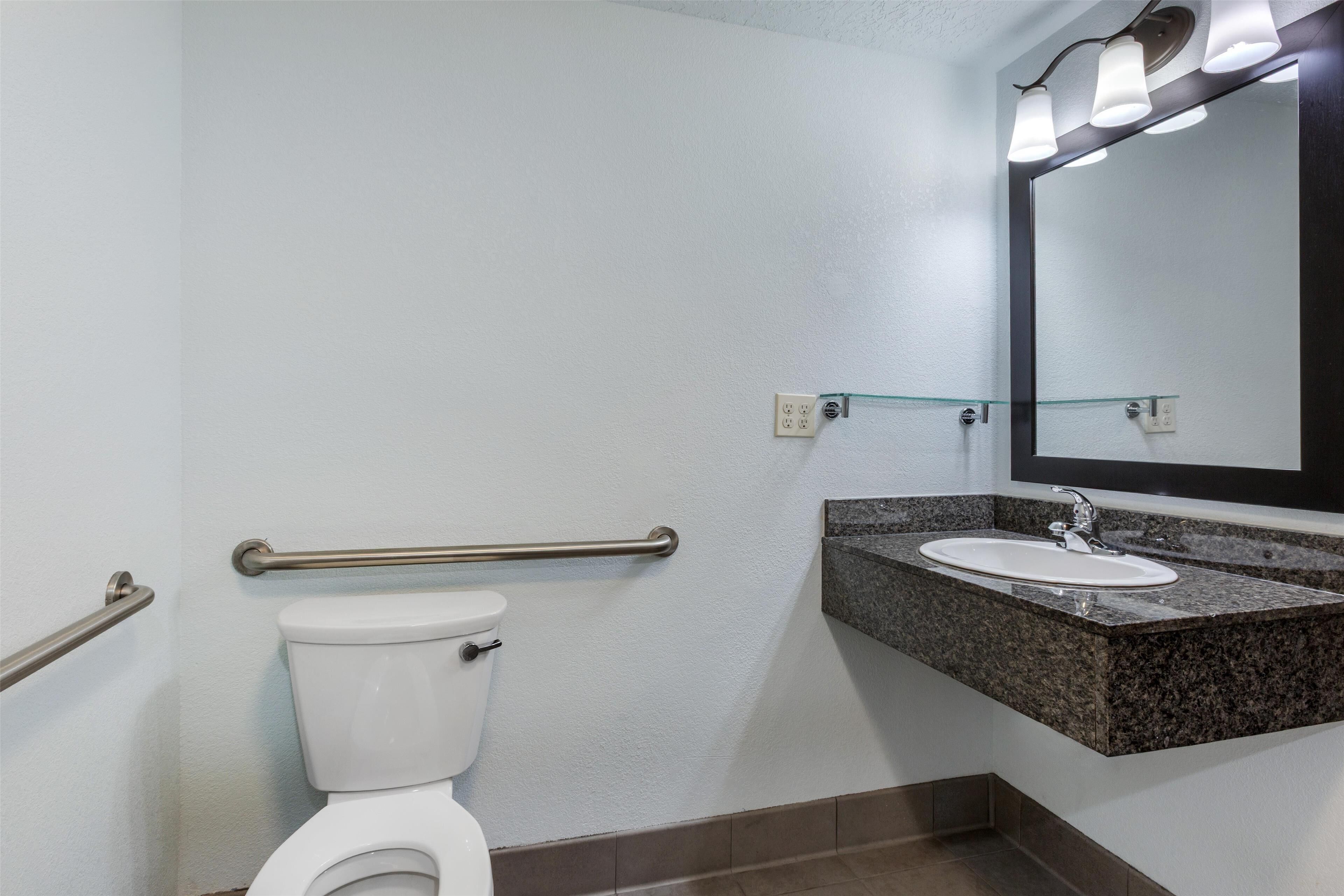 Un baño con inodoro blanco, lavamanos de granito, espejo y barandilla.