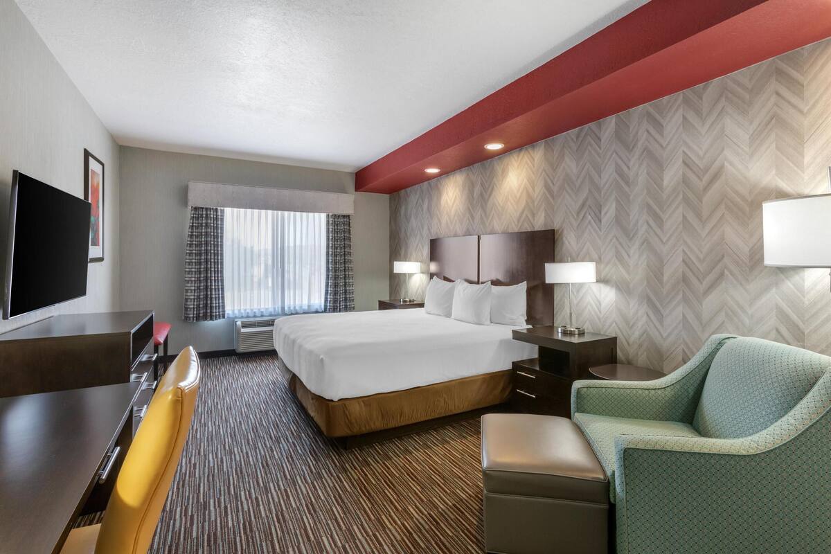A hotel room with a large bed, a desk with a chair, a TV, and a seating area with a chair and ottoman.