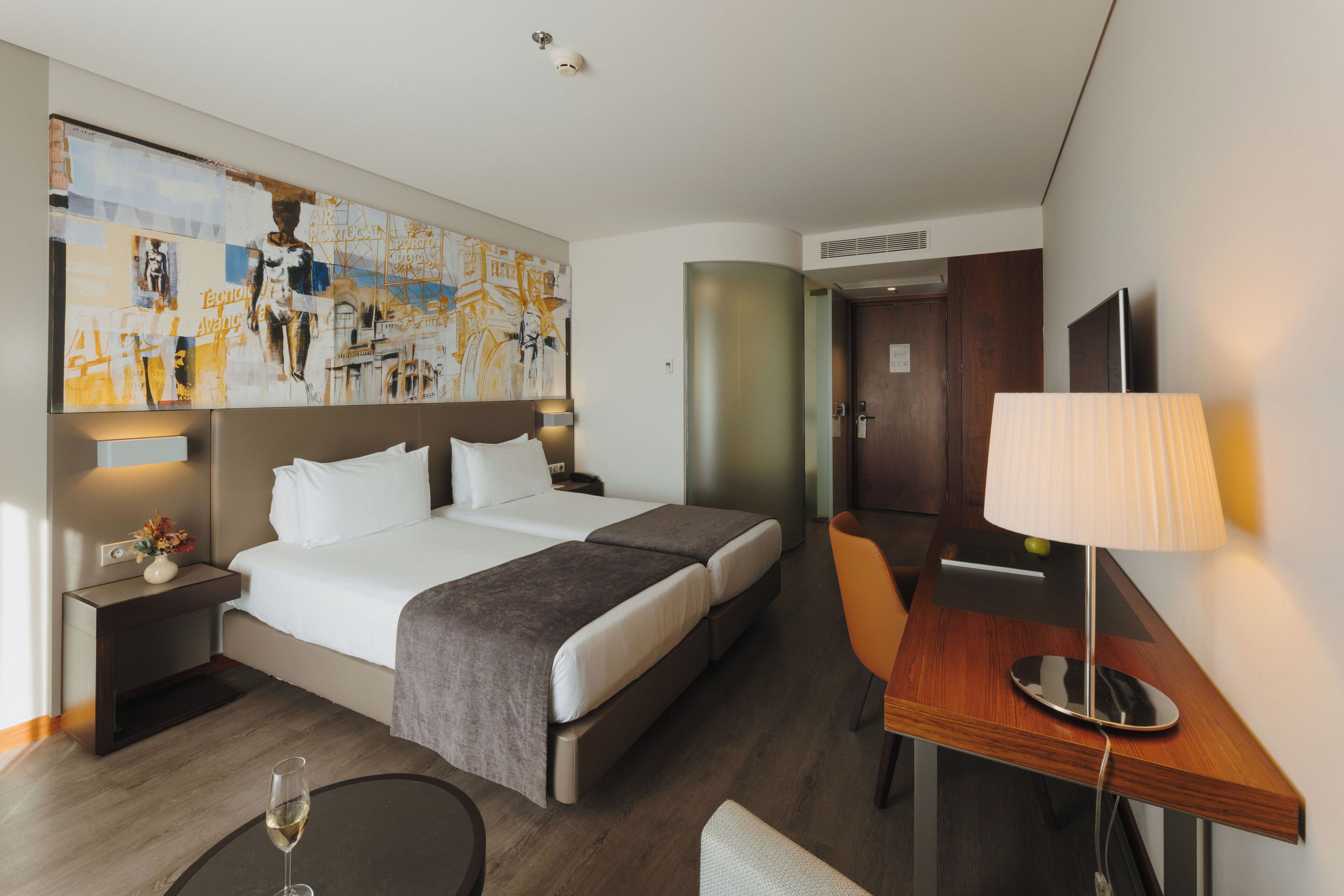 Una habitación de hotel con una cama grande, un escritorio con una lámpara, una silla, una mesita con una copa de vino y una pared decorada con obras de arte.