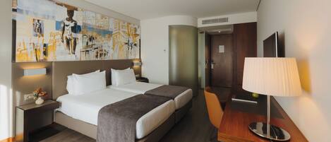 Una habitación de hotel con una cama grande, un escritorio con una lámpara, una silla, una mesita con una copa de vino y una pared decorada con obras de arte.