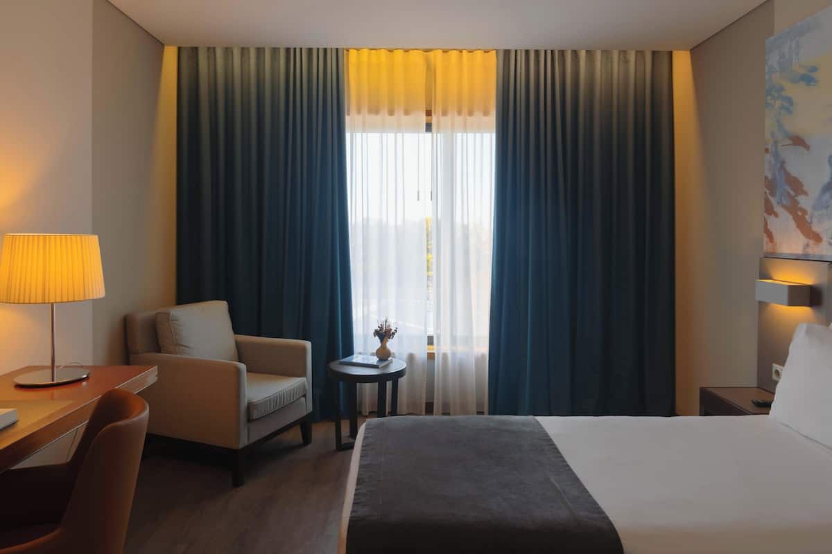 A hotel room with a bed, a desk, a chair, a small table with a vase, and a painting on the wall.