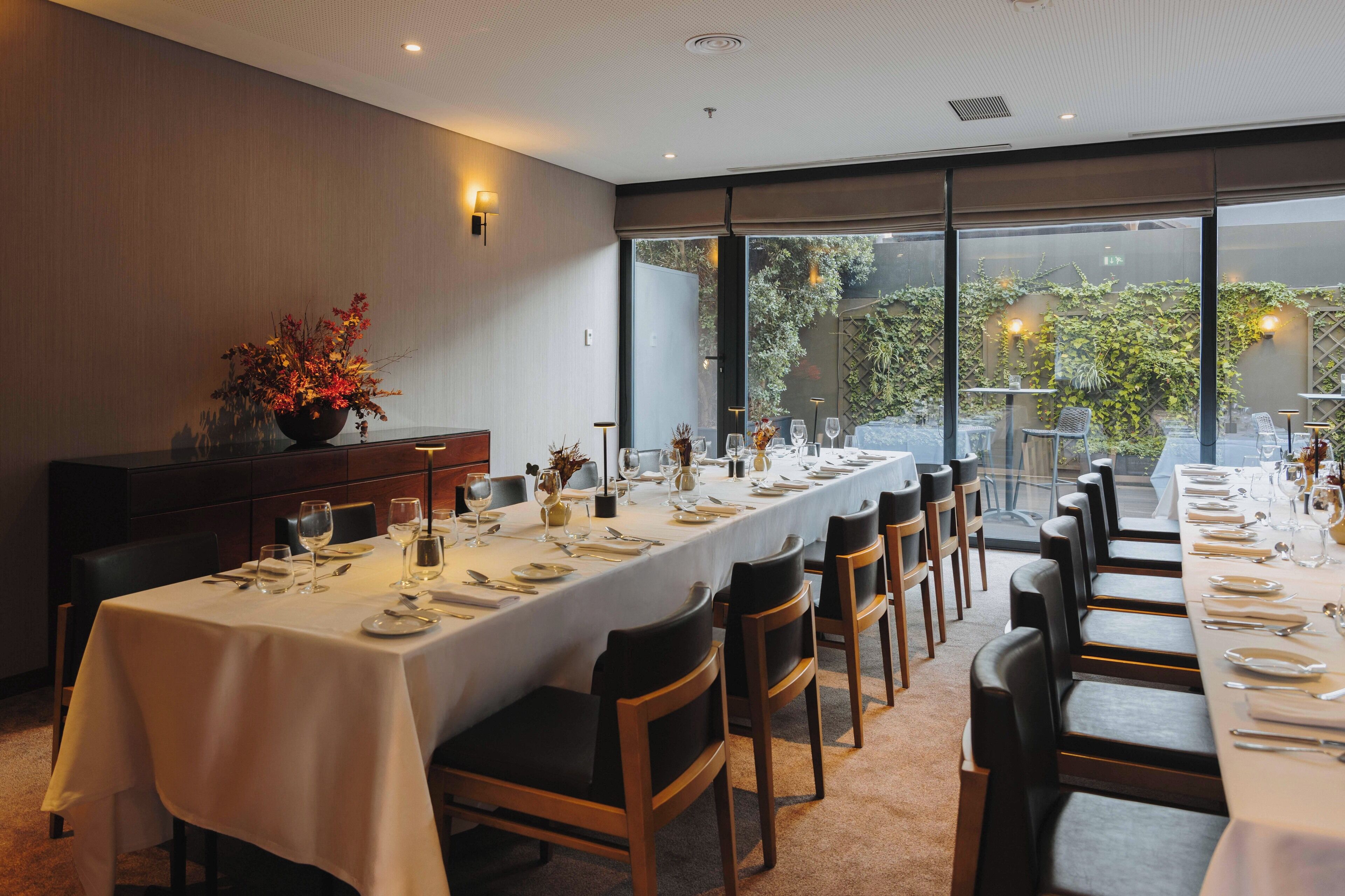 A long dining table set for a meal with chairs, a vase of flowers, and a view of the outdoors through large windows.