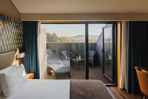 Habitación de hotel con cama, balcón con sillas de mimbre, una mesa con una botella y vistas de la ciudad.