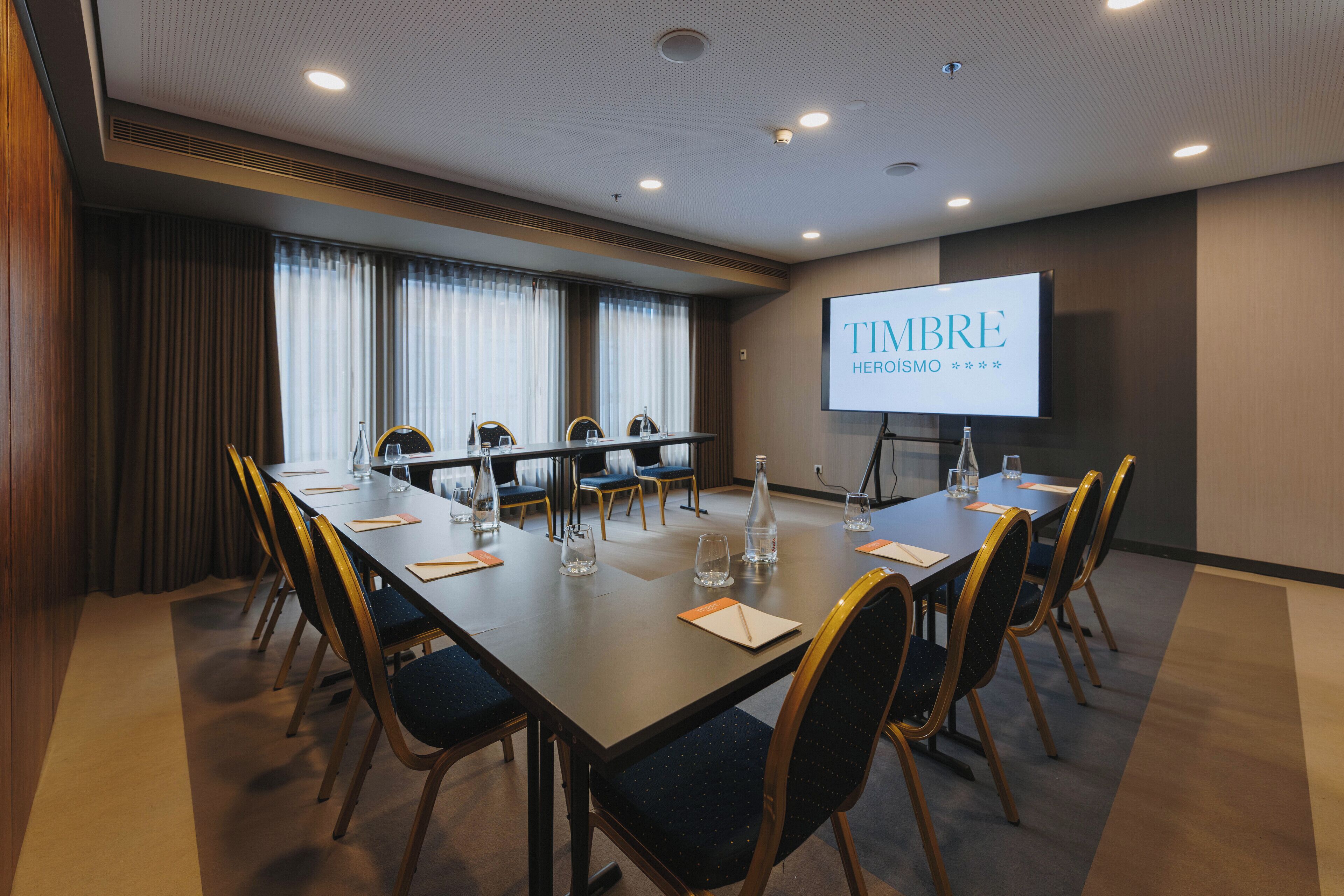 A conference room with a long table, chairs, and a projection screen displaying "Timbre Heroismo".
