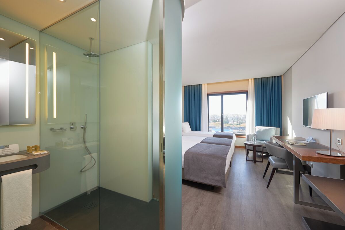 Habitación de hotel con ducha de vidrio, cama, escritorio con silla, televisor y ventana con cortinas.