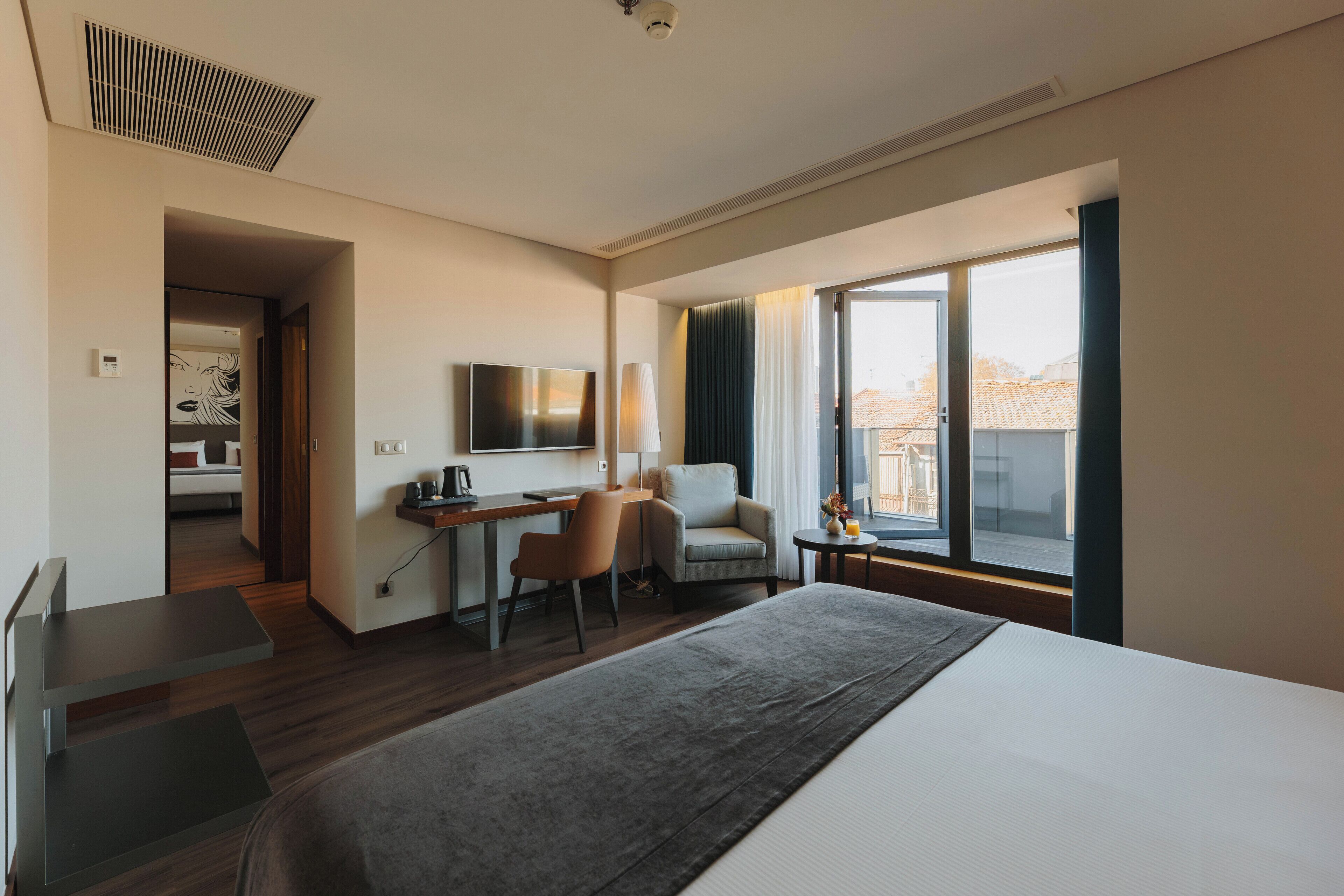Habitació d'hotel moderna amb un llit gran, una taula de treball amb una cadira, una televisió i una finestra amb cortines.