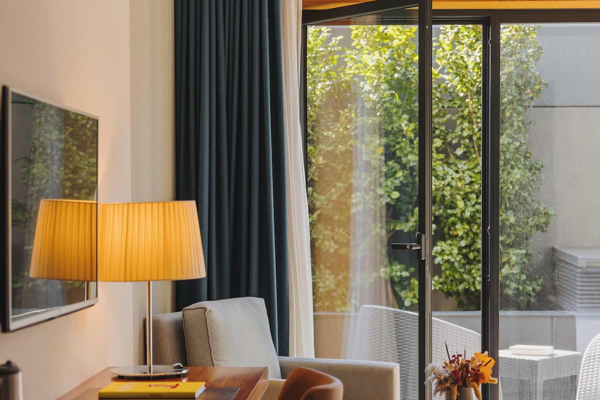 A hotel room with a desk, two chairs, a sofa, a small table with a vase, and a large window with curtains.