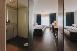 Habitación de hotel con ducha de vidrio, cama con cabecero de madera, escritorio con silla y ventana con cortinas.