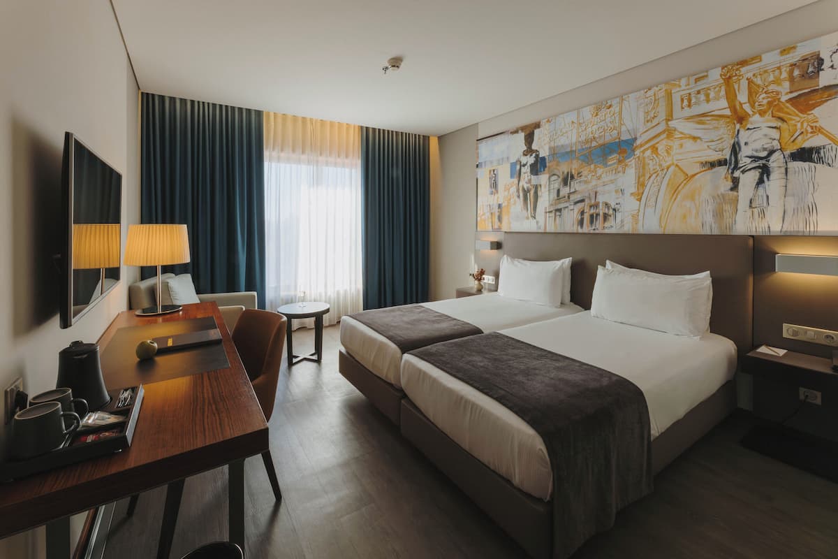 A hotel room with two beds, a desk, a chair, a small table, and a large painting on the wall.