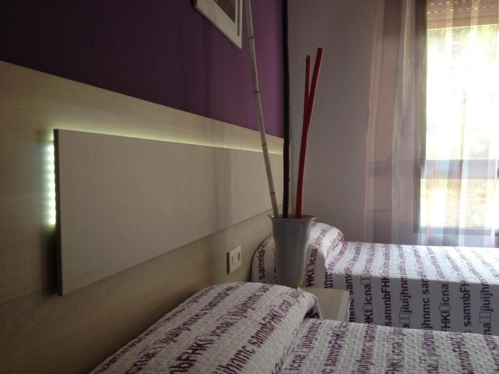 A room with two beds, a headboard, a vase with red and white sticks, and a window with sheer curtains.