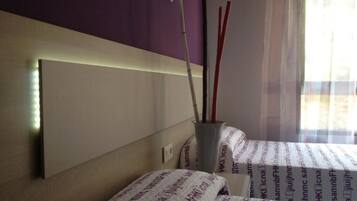 A room with two beds, a headboard, a vase with red and white sticks, and a window with sheer curtains.