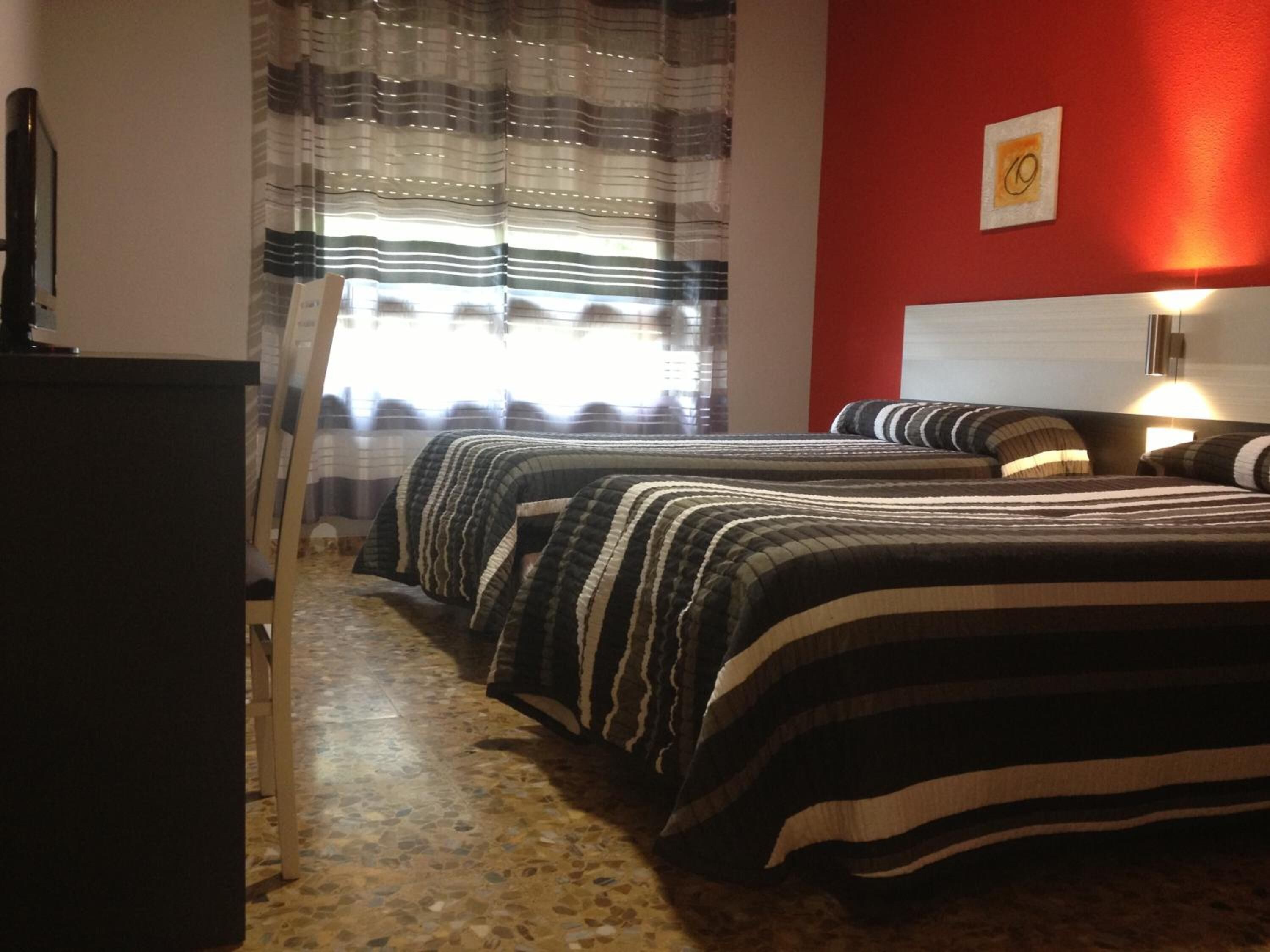 Une chambre d’hôtel avec deux lits, une literie à rayures, un mur d’accent rouge, une fenêtre avec des rideaux et un bureau avec une lampe.