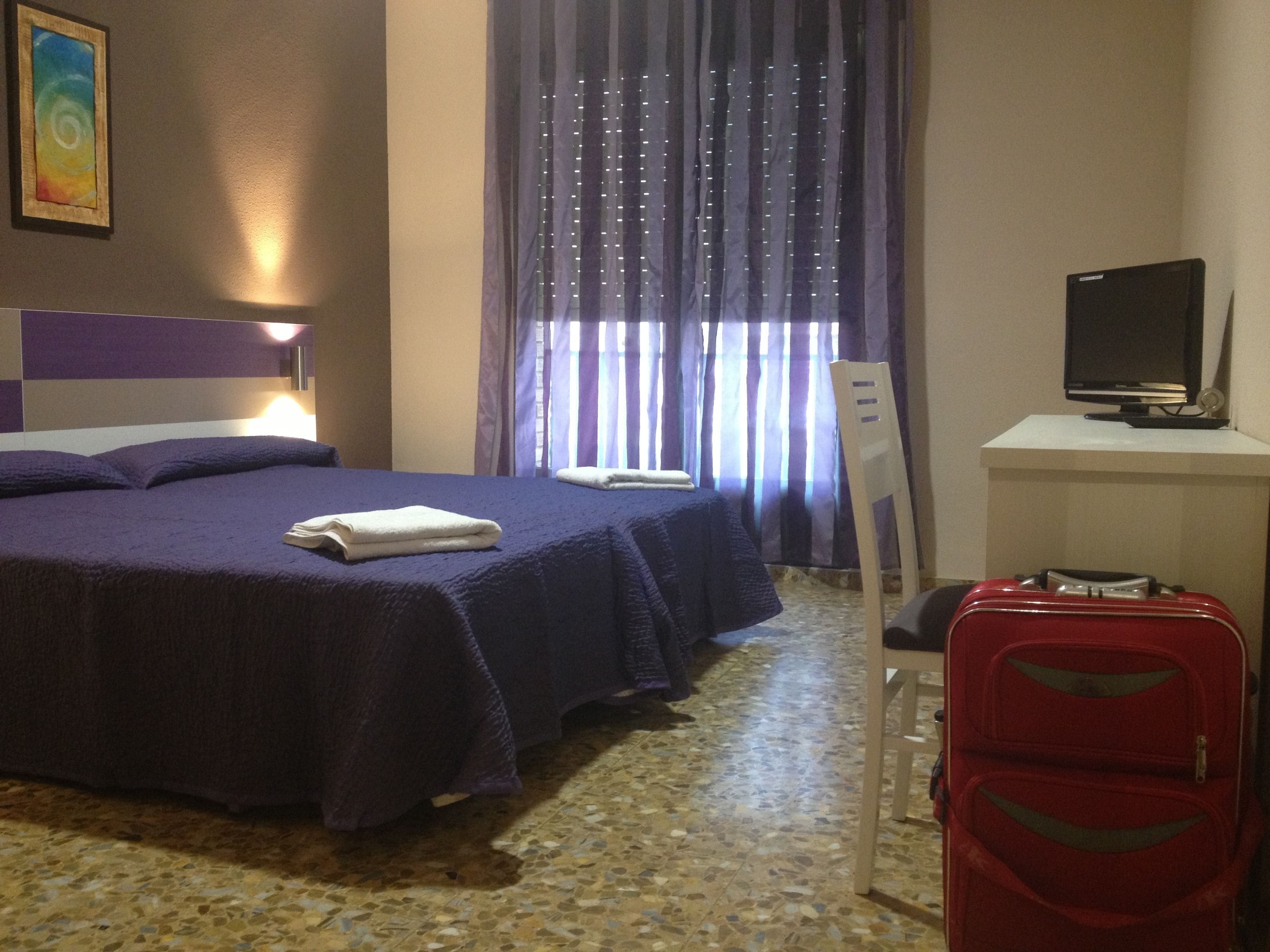 Quarto de hotel com uma cama, uma mesa de trabalho com televisão, uma mala vermelha e uma janela com cortinas.