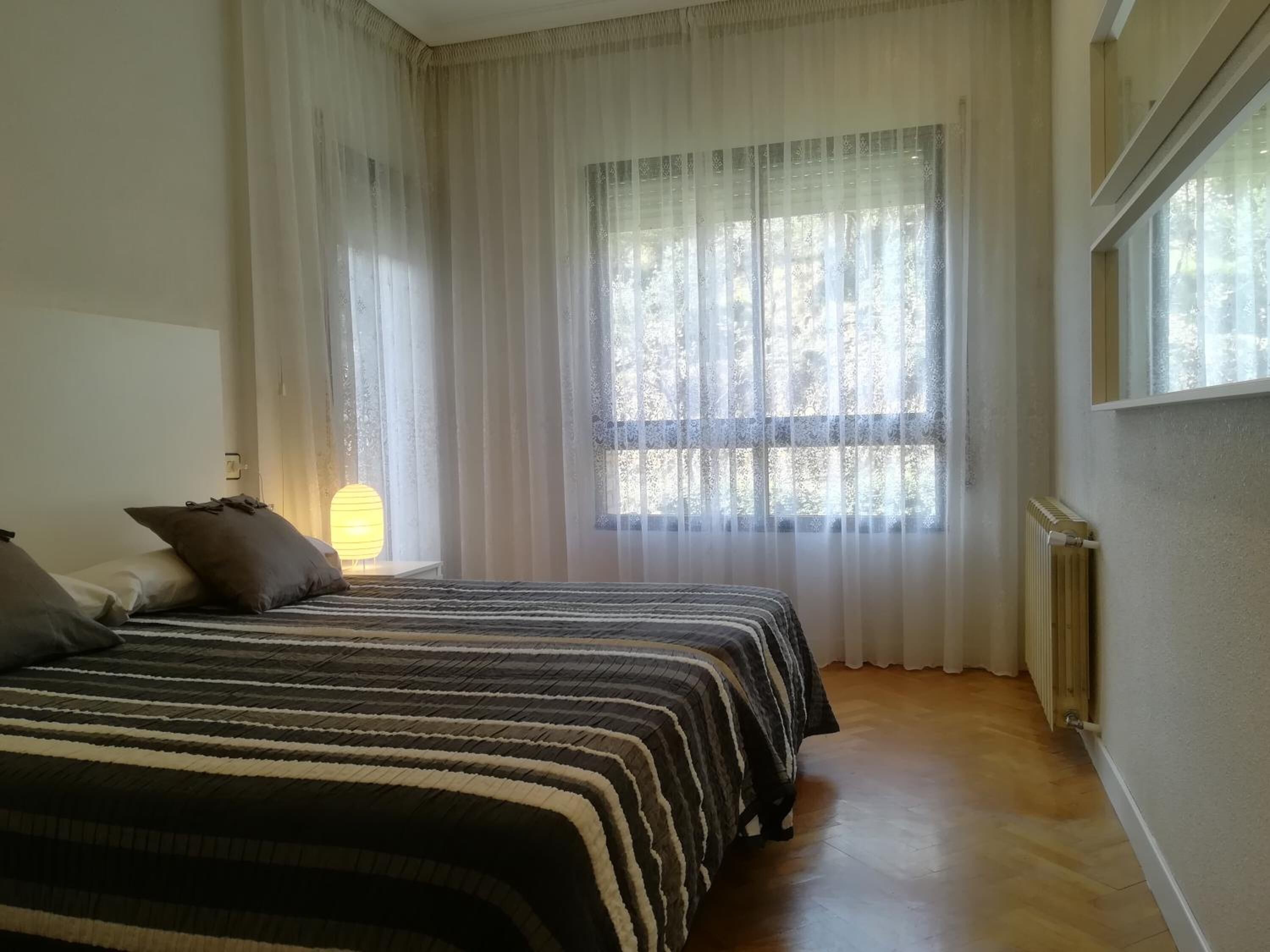 Un dormitorio con dos camas, cortinas translúcidas, un radiador y una ventana con vistas a árboles.