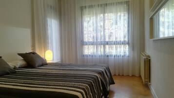 Un dormitorio con dos camas, cortinas translúcidas, un radiador y una ventana con vistas a árboles.