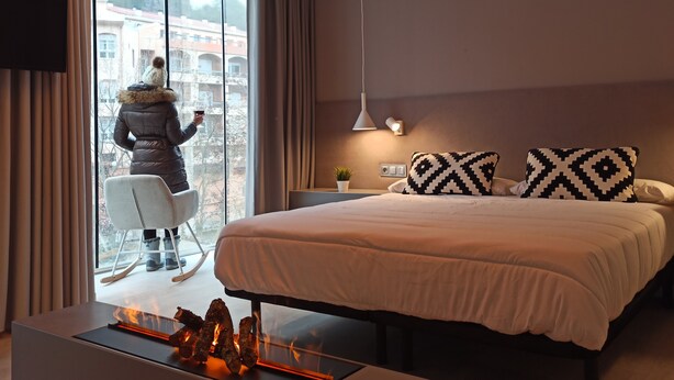 A modern hotel room with a large bed, a seating area with a chair, a fireplace, and a view of buildings through large windows. - Hotel Marrodan (Arnedillo)