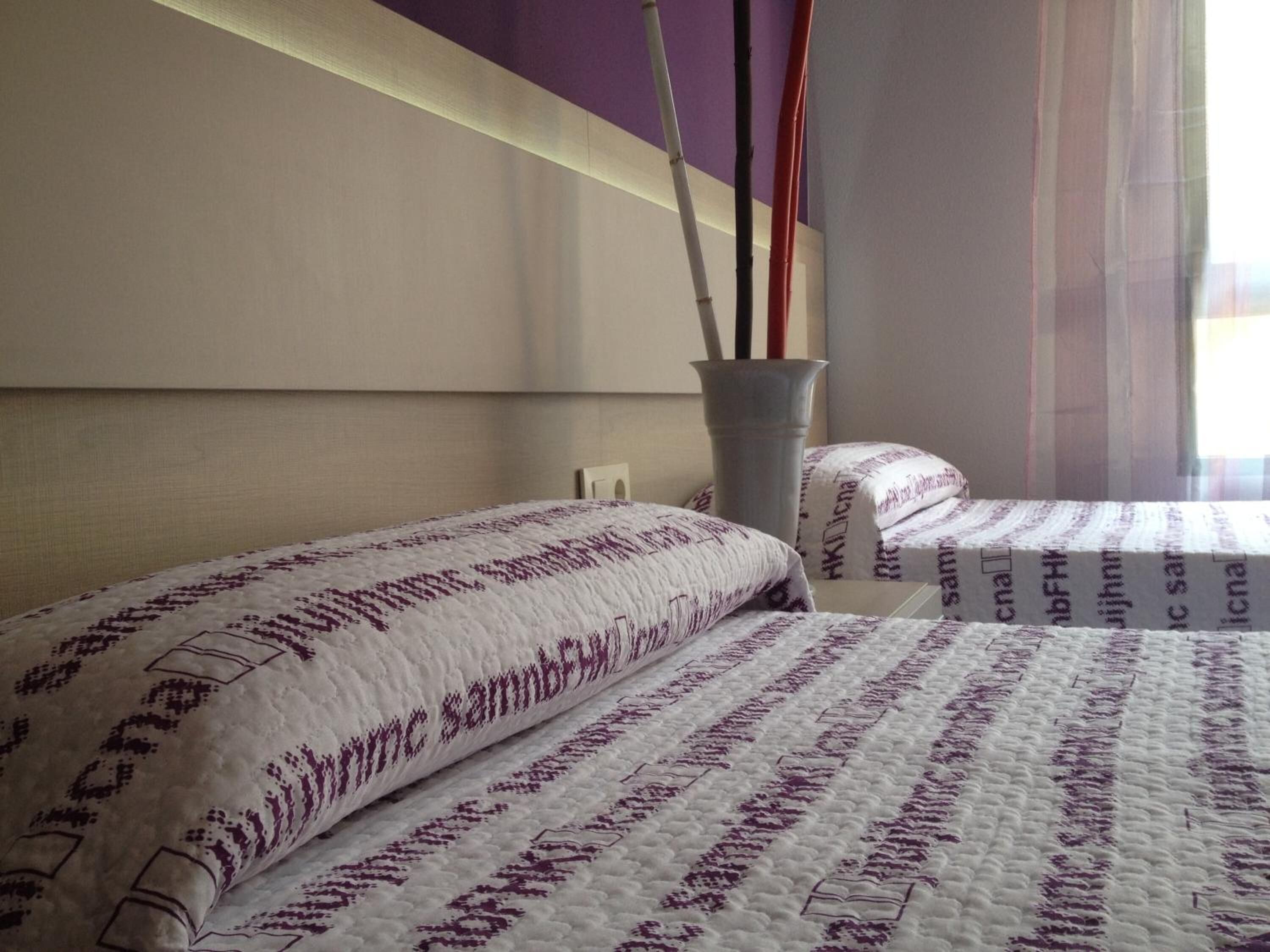Une chambre d’hôtel avec deux lits, un vase décoré de branches et une fenêtre ornée de rideaux légers.