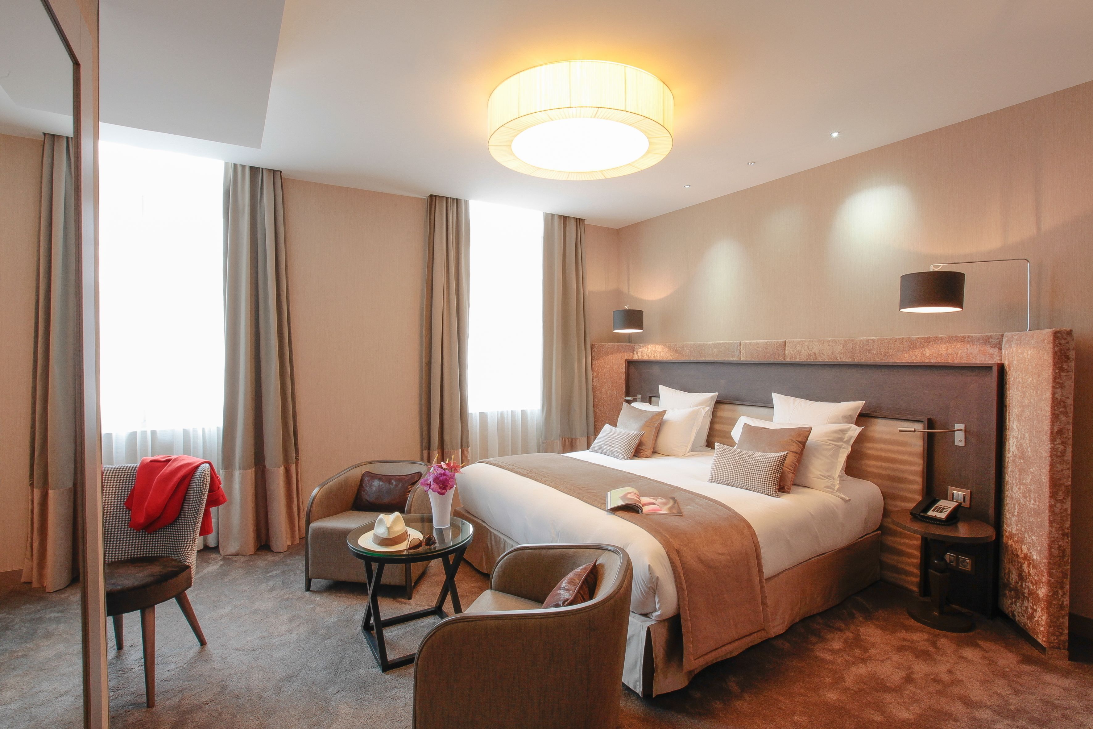 A hotel room with a large bed, a desk, two chairs, a small table with a vase and flowers, and a nightstand with a lamp.